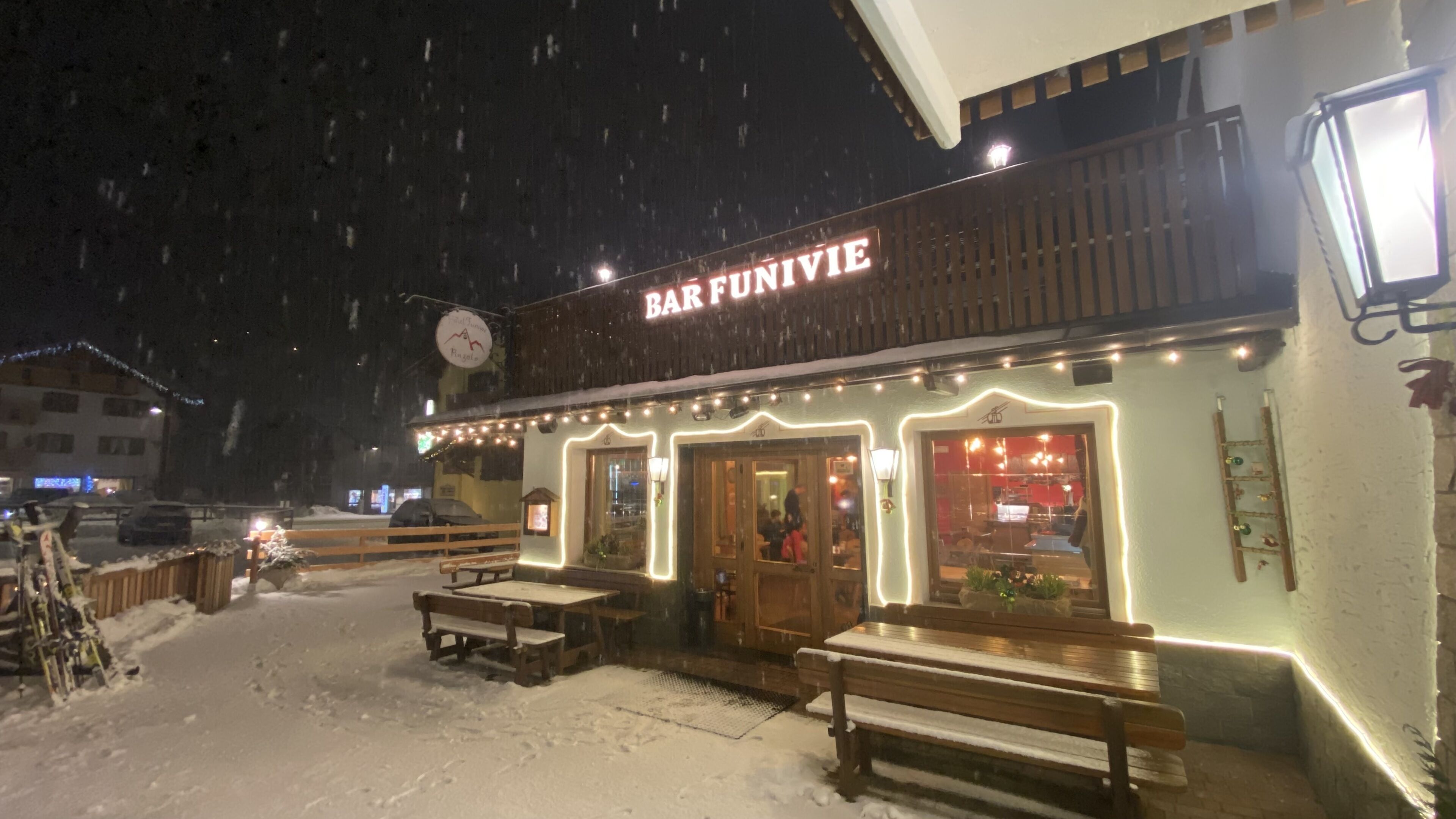 A snow-covered outdoor seating area with wooden benches and tables in front of a bar named "BAR FUNIVIE".