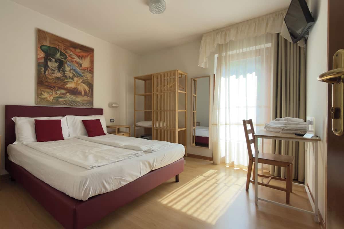 A hotel room with a bed, a desk with a chair, a wardrobe, and a painting on the wall.