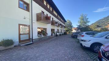 A building with a white facade, wooden balconies, and a paved courtyard with parked cars.