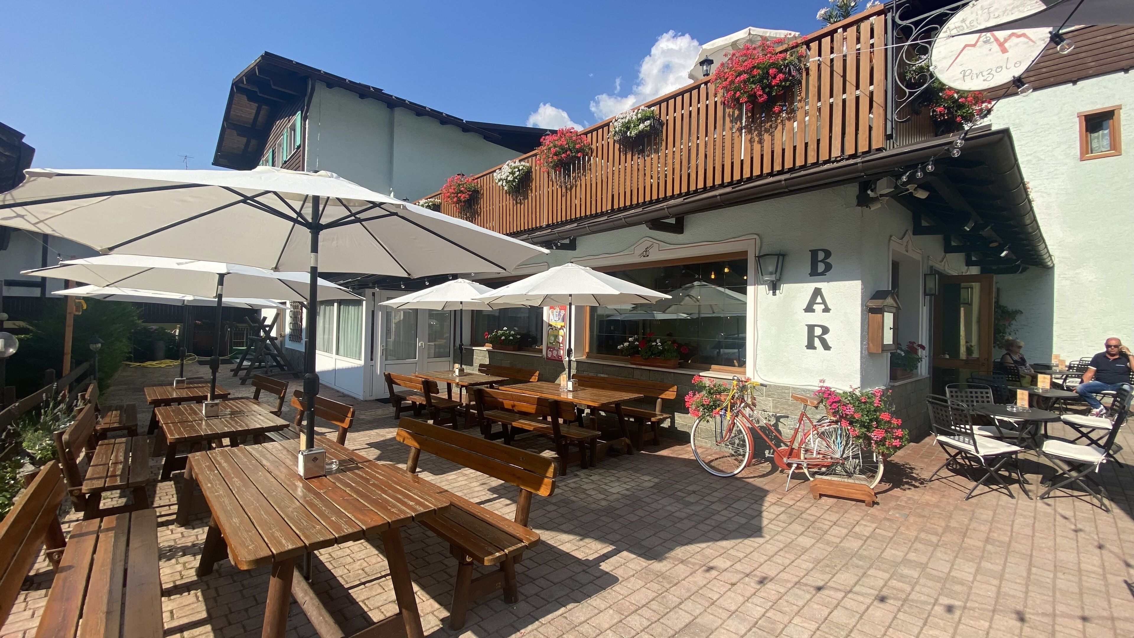 Ein Außenbereich mit Holztischen und weißen Sonnenschirmen, eine Bar mit der Aufschrift 'BAR' und Blumenkörben am Balkon.