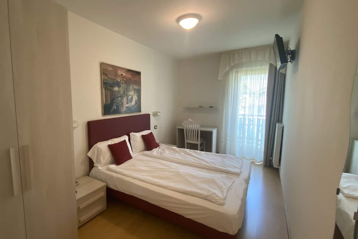 Un lit double avec du linge de lit blanc et des oreillers rouges, une table de chevet, un petit bureau avec une chaise, un tableau au mur et une fenêtre avec des rideaux.