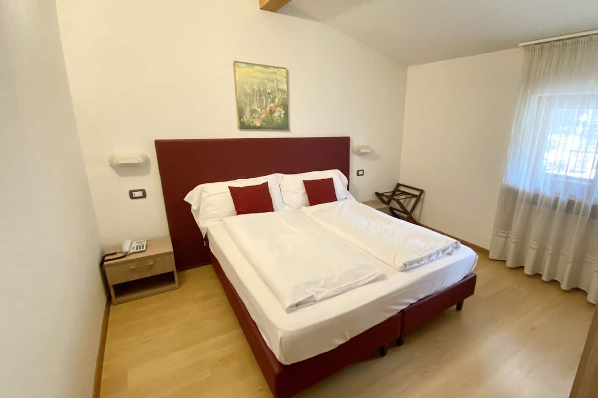 Un lit bien fait, avec du linge de lit blanc et des oreillers rouges, dans une pièce au plancher en bois, dotée d’une tête de lit rouge et ornée d’un tableau au mur.