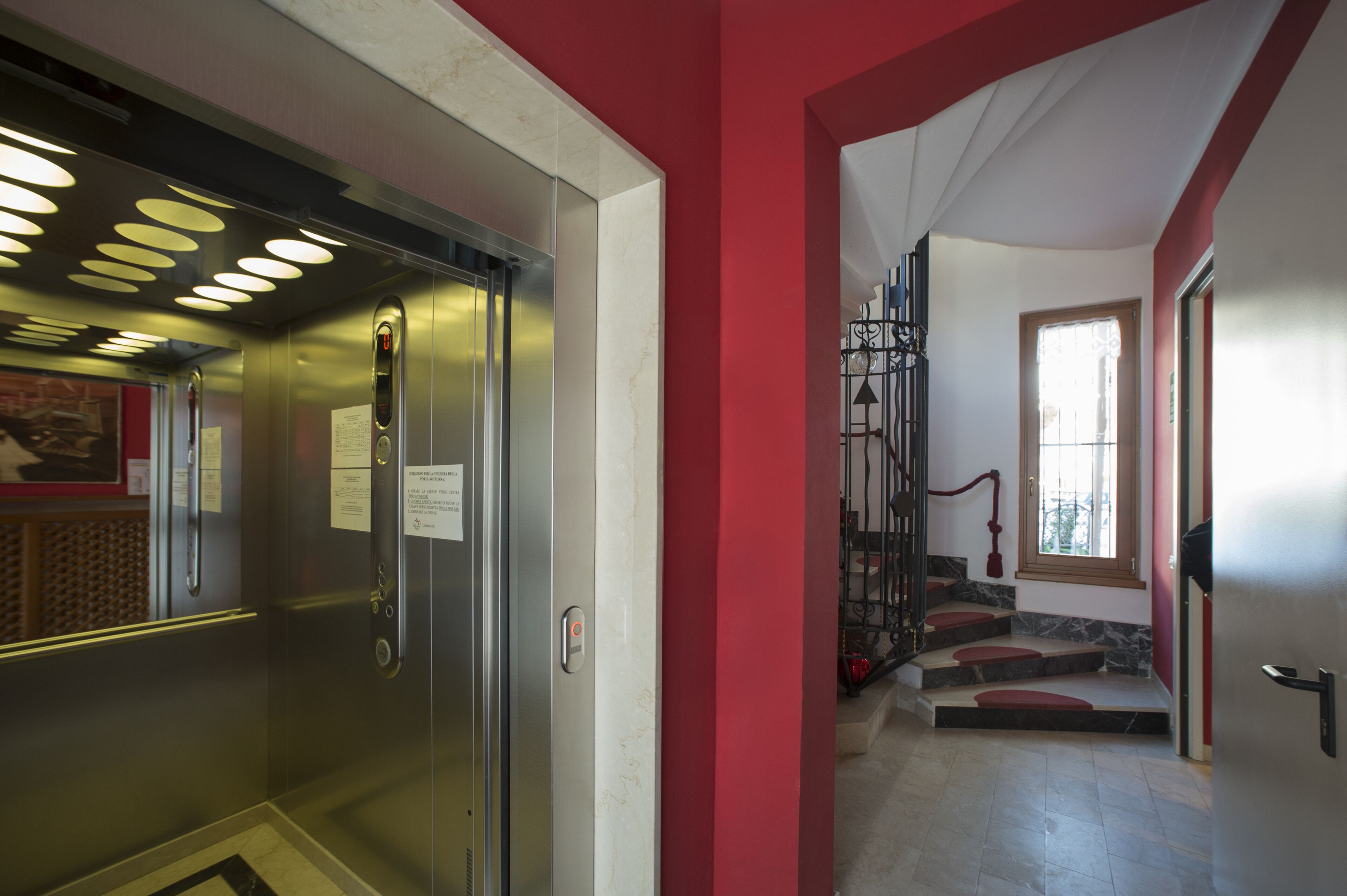 Un ascenseur aux finitions métalliques, un mur rouge et un escalier avec des marches rouges menant à une porte dotée d’une fenêtre.