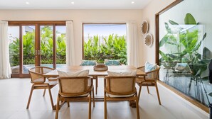 Comedor con mesa de madera y sillas de mimbre, ventanas amplias con vistas a zonas verdes y un panel de vidrio que refleja plantas.