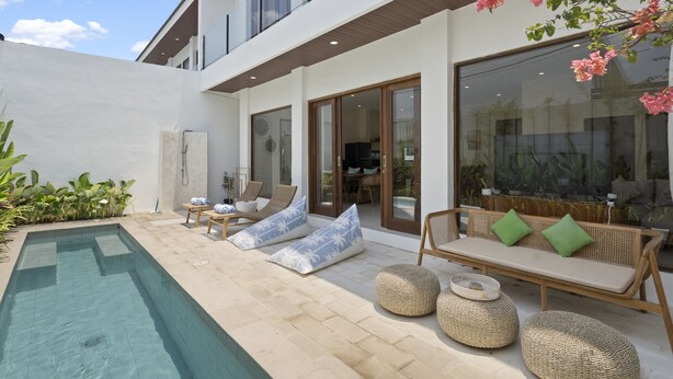 Un área de piscina moderna al aire libre con sillones de descanso y un sofá, rodeada por un edificio blanco con grandes ventanales.
