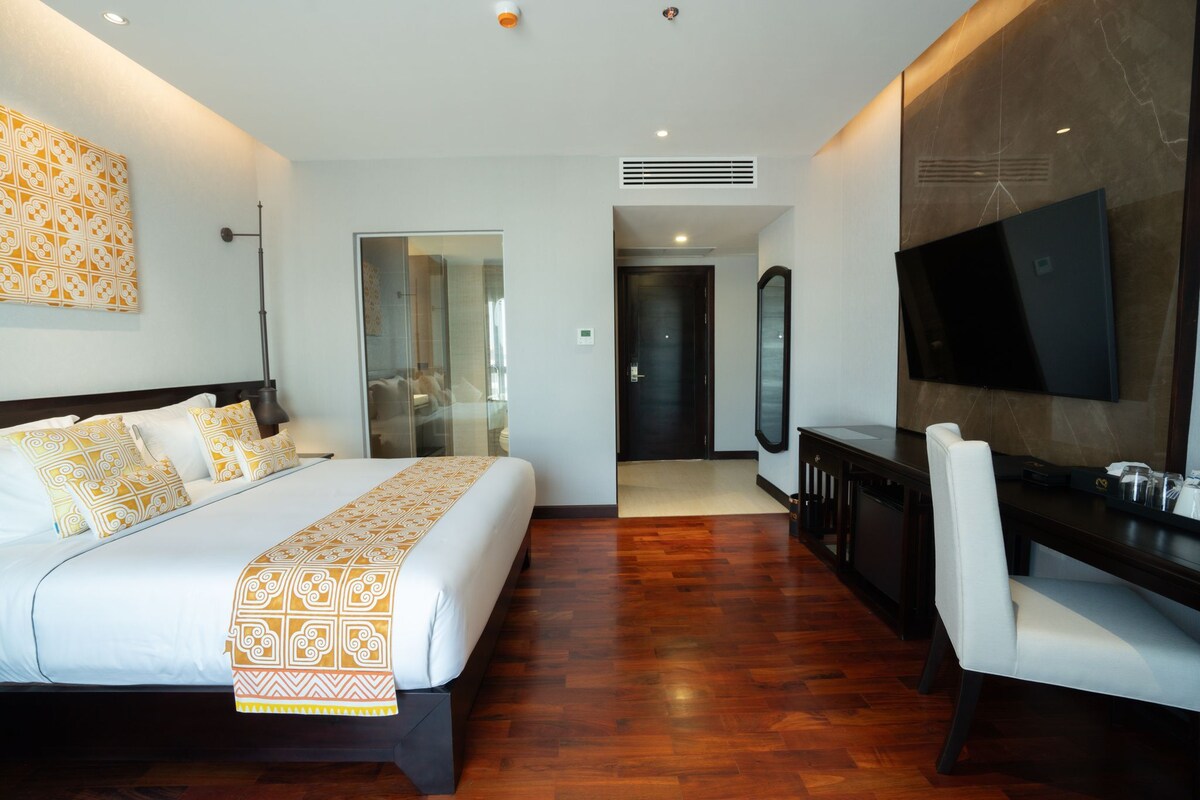 A modern hotel room with a large bed, a desk, a chair, a TV, and a bathroom visible through a glass door.