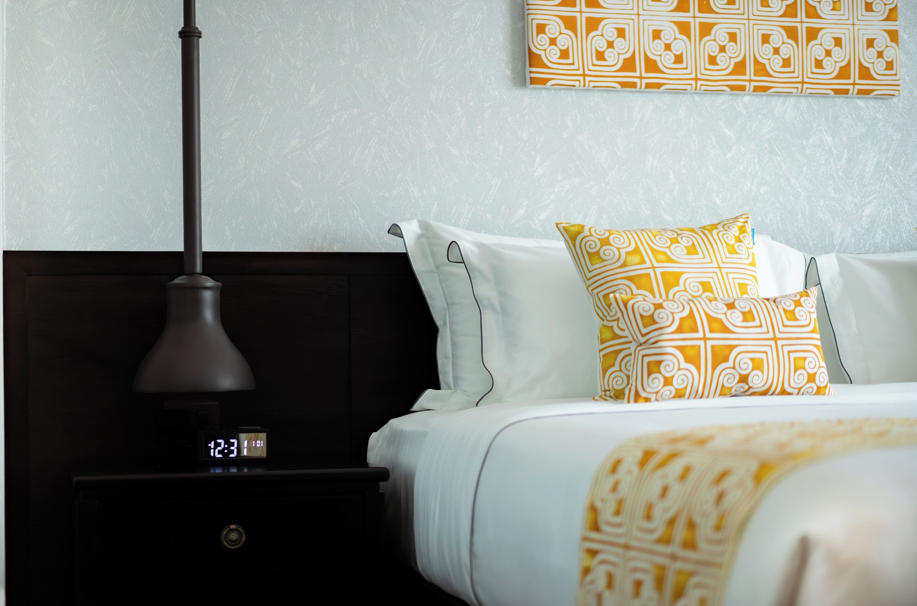 Una cama bien hecha con almohadones blancos y amarillos estampados, una mesita de noche oscura con un reloj digital, y una pared con una obra de arte de diseño geométrico.
