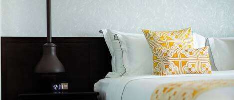 Una cama bien hecha con almohadones blancos y amarillos estampados, una mesita de noche oscura con un reloj digital, y una pared con una obra de arte de diseño geométrico.