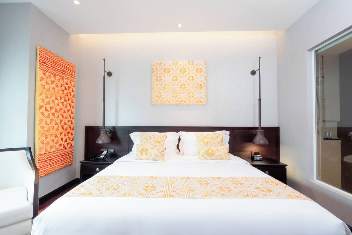 Une chambre d’hôtel moderne dotée d’un grand lit, de deux tables de chevet, d’une petite table, d’une œuvre d’art accrochée au mur et d’une fenêtre offrant une vue agréable.