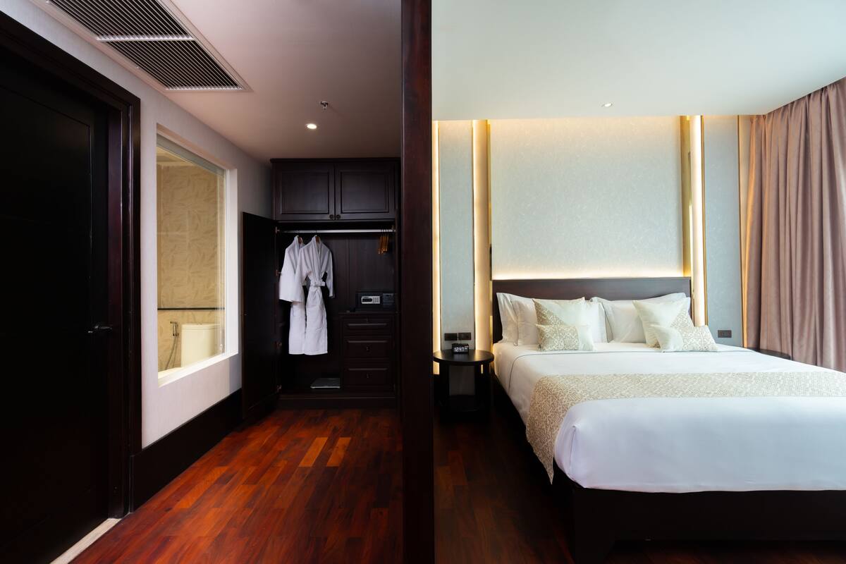 Une chambre d’hôtel avec un plancher en bois, un lit avec une literie blanche, une table de chevet, une armoire et une salle de bain visible à travers une porte ouverte.