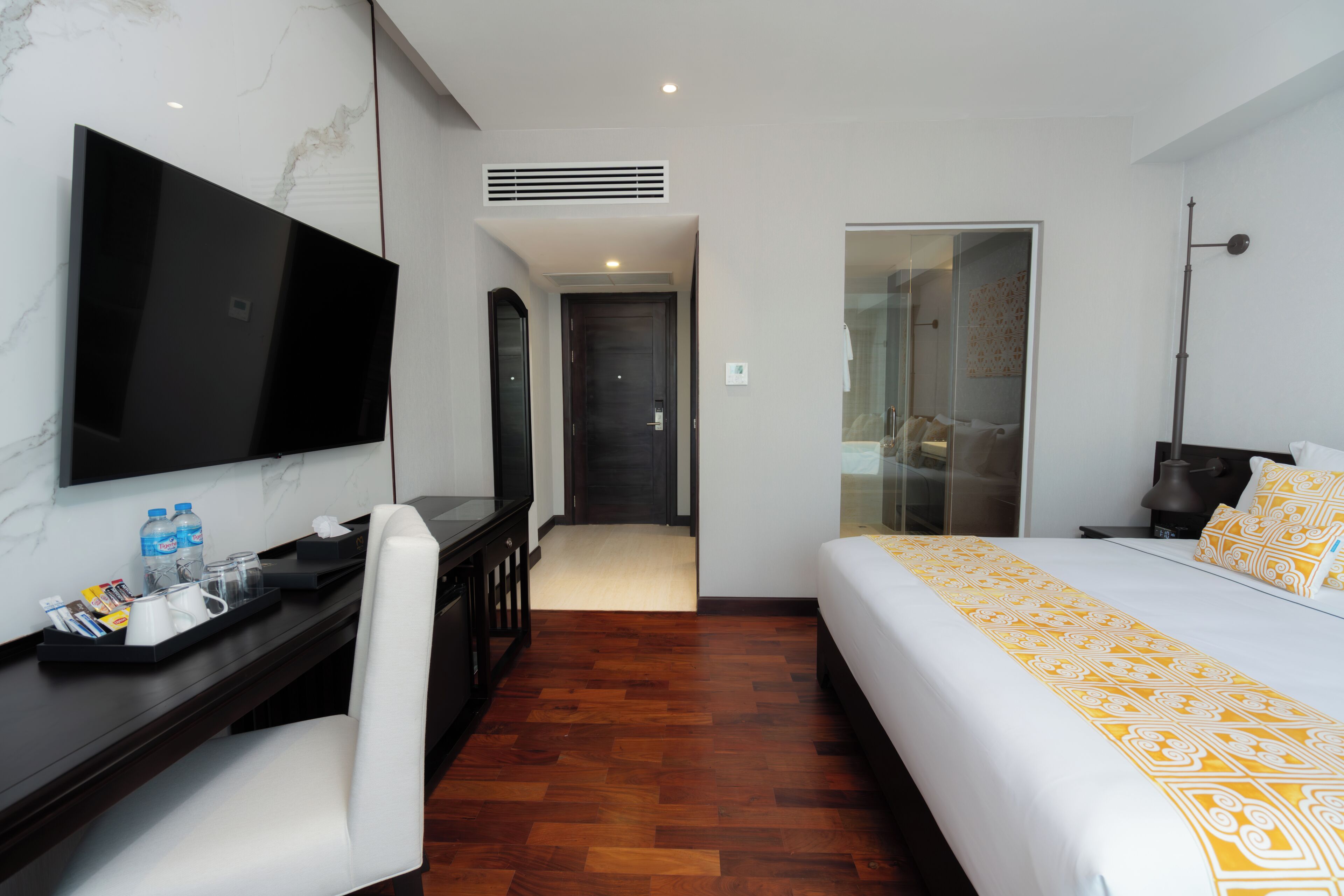 A hotel room with a bed, a desk with a flat-screen TV, a chair, a coffee tray, and a bathroom visible through a glass door.