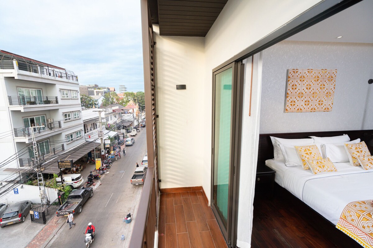 A balcony with a bed, a view of a busy street, and a building with multiple floors.