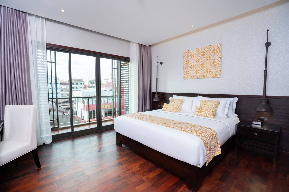 Une chambre d’hôtel moderne, dotée d’un grand lit, d’une chaise, d’une table de chevet et offrant une vue sur la ville grâce à de grandes fenêtres.