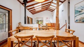 A dining area with a large wooden table and chairs, a high ceiling with exposed beams, a chimney, and a painting on the wall.