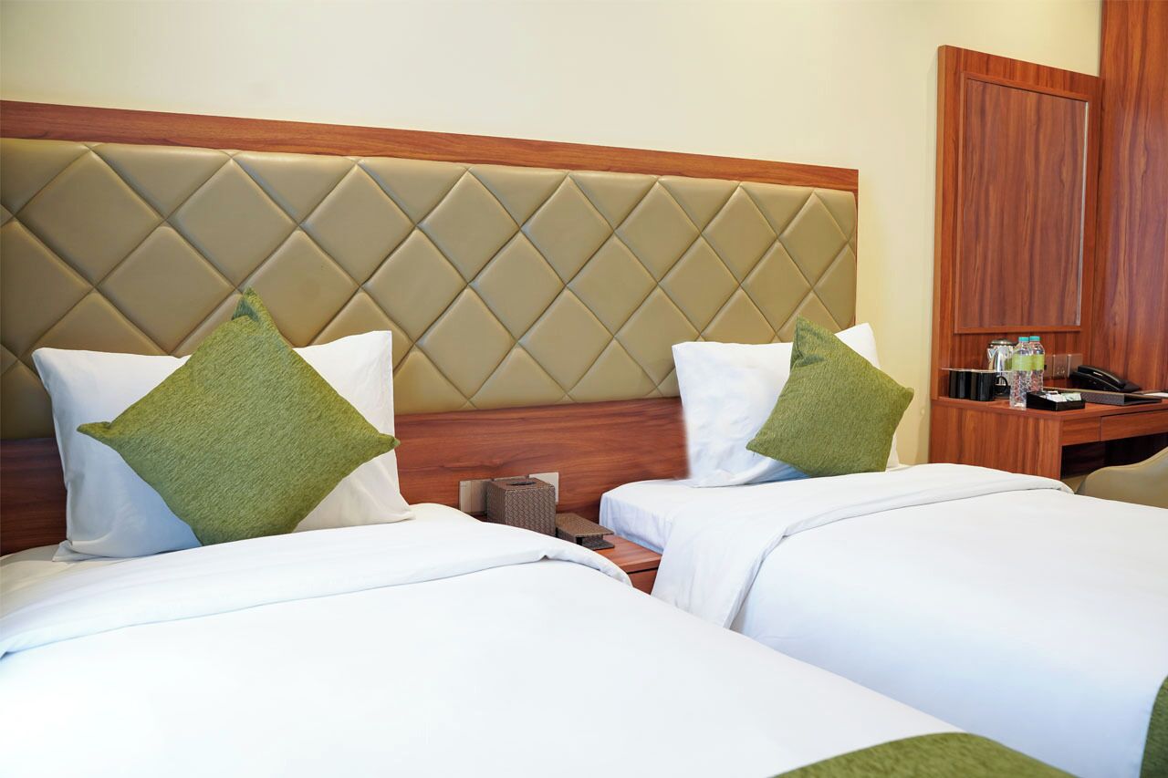 Habitación de hotel con dos camas, cada una con ropa blanca y almohadas verdes. El cabecero está capitonado y con paneles de madera. Hay un escritorio de madera con un teléfono y una mesita con una botella y una caja de pañuelos.
