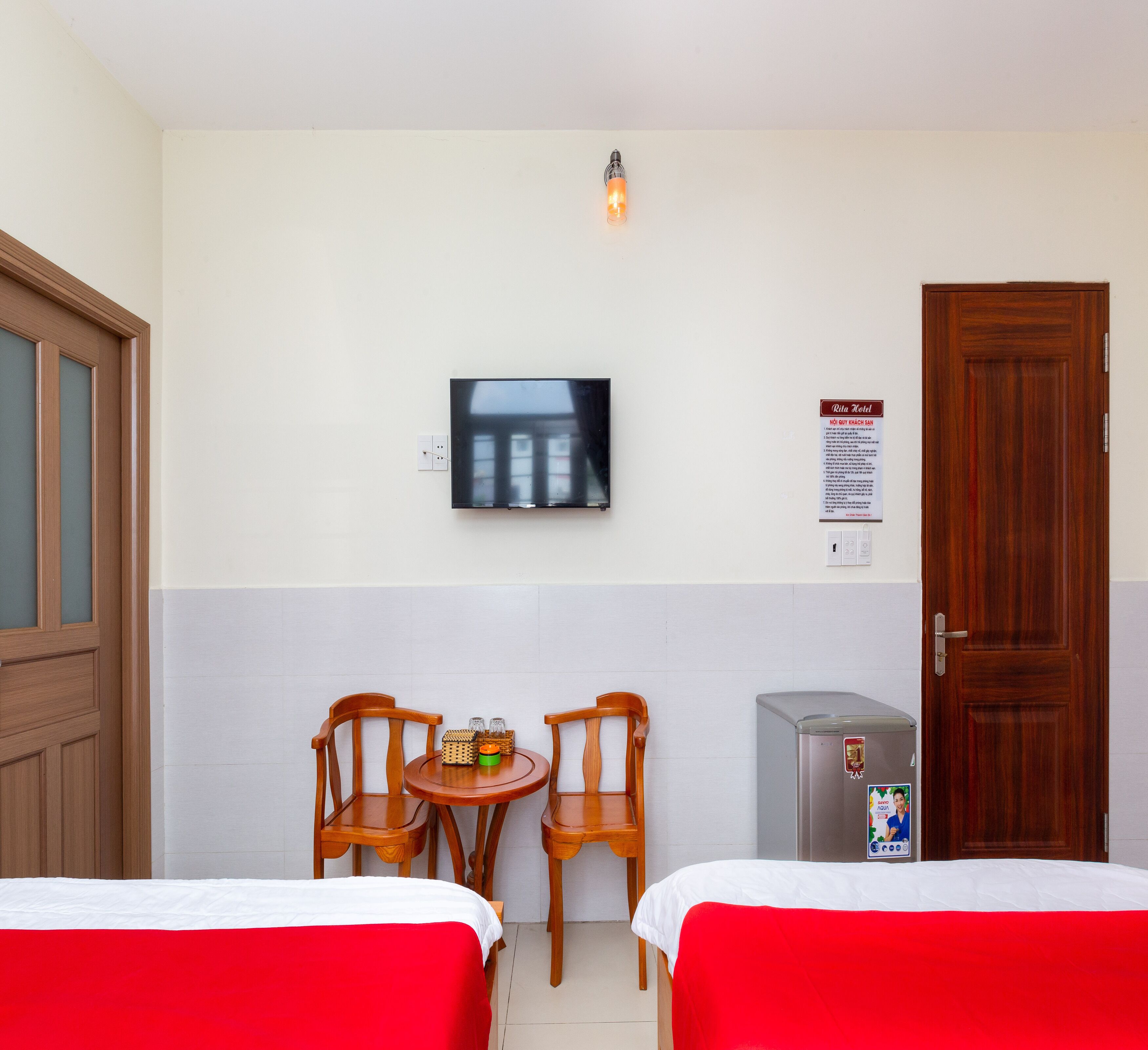 A hotel room with two beds, a TV mounted on the wall, a small table with chairs, a refrigerator, and a door with a sign.