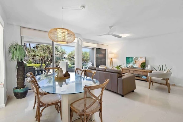 A modern living room with a glass dining table, wicker chairs, a sofa, and a television.