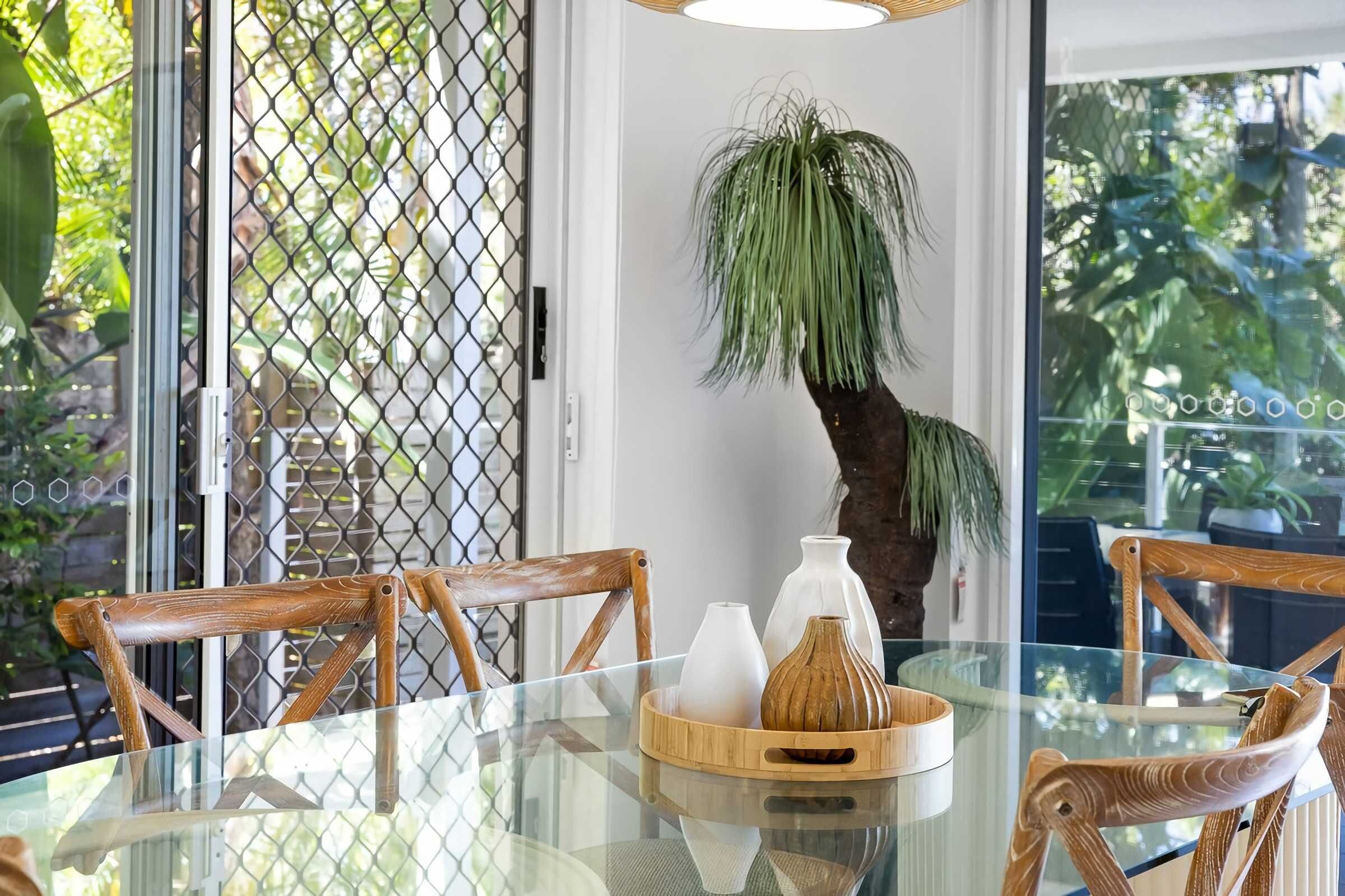 Een eettafel met glazen blad, houten stoelen, een decoratieve plant en een dienblad met vazen.