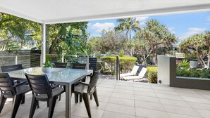 Un espace extérieur abrité, aménagé pour les repas, avec une table et des chaises en verre, entouré d’une végétation luxuriante et de palmiers.