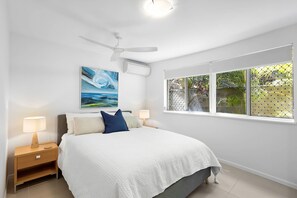 A bedroom with a bed, two bedside tables, a ceiling fan, a window with a view of greenery, and a painting on the wall. - The Sunshine Villa 2 Min walk to Village Beach (Noosa)