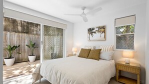 Une chambre à coucher comprenant un lit, deux tables de chevet avec des lampes, un ventilateur de plafond, une fenêtre avec vue et une œuvre d’art accrochée au mur.