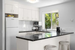 A modern kitchen with white cabinets, a black countertop, a microwave, and a window with a view of greenery. - The Sunshine Villa 2 Min walk to Village Beach (Noosa)