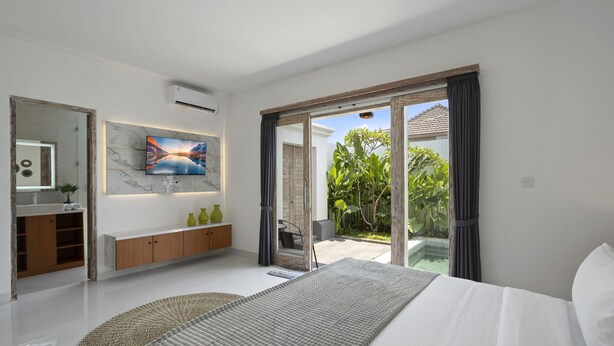 Quarto moderno com cama grande, televisão na parede, banheiro com toucador e porta de correr de vidro para um pátio.