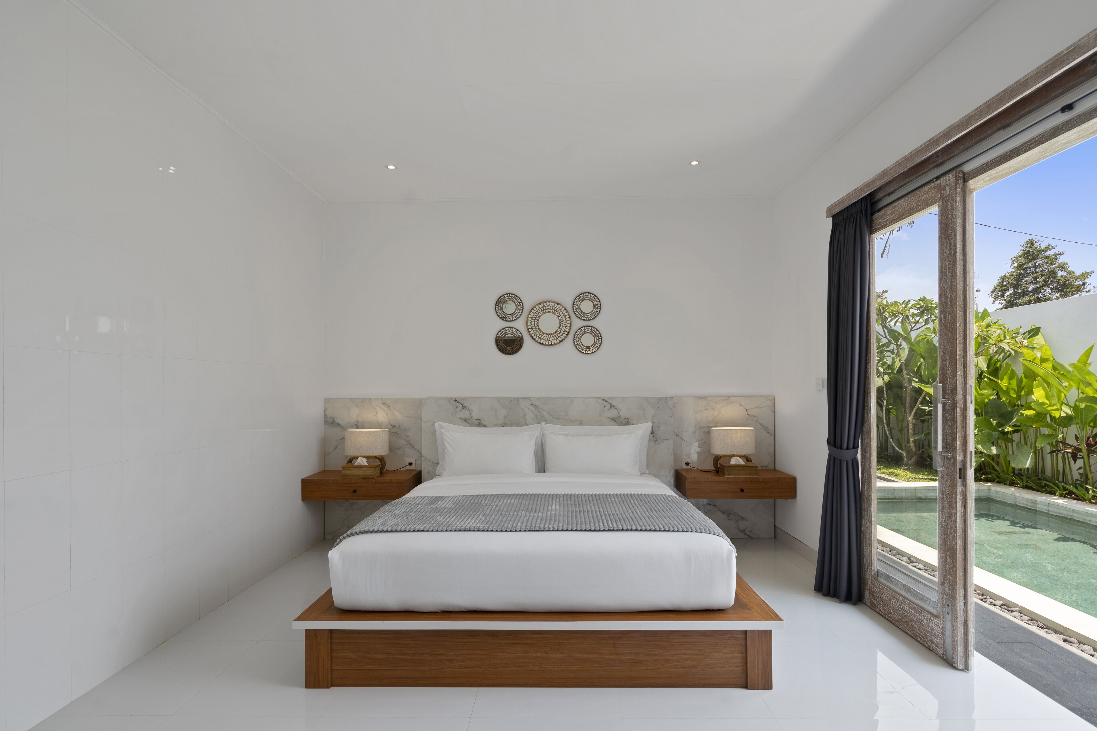 Une chambre moderne avec un grand lit, une tête de lit en marbre, deux tables de chevet avec des lampes et une vue sur une piscine, visible à travers une porte vitrée.