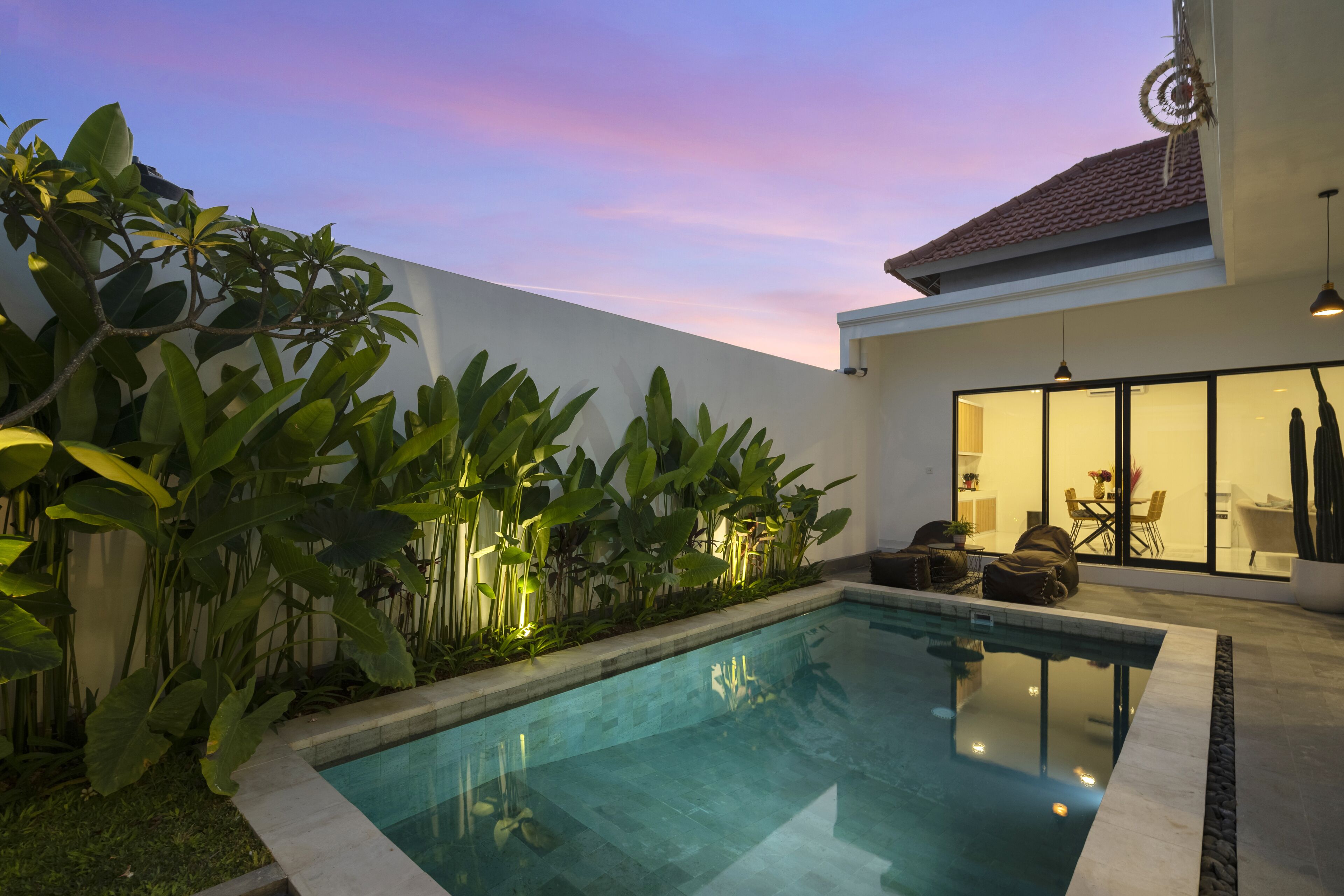 A modern outdoor pool area with a clear blue pool, surrounded by lush greenery and a white wall.