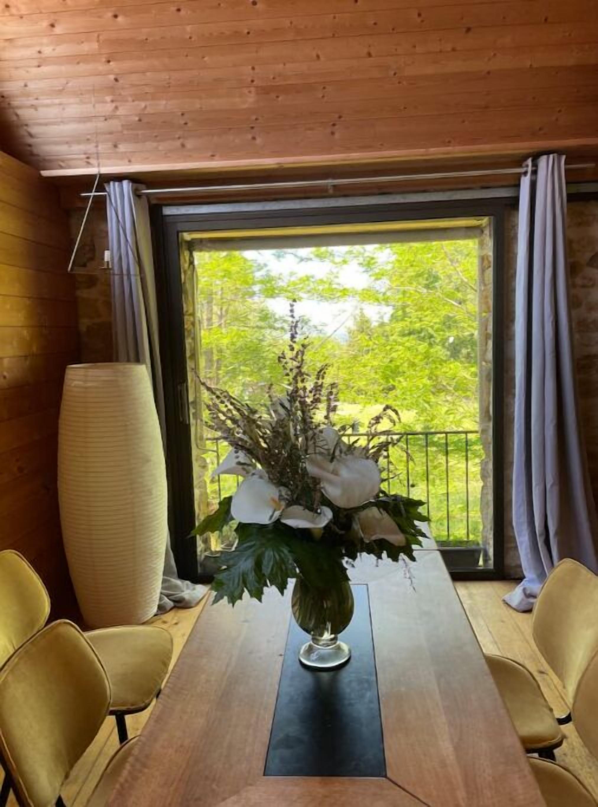 Une table à manger en bois, ornée d’un vase de fleurs au centre, et flanquée de deux chaises, dans une pièce aux murs en bois et dotée d’une grande fenêtre avec des rideaux.