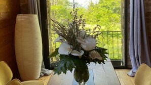Una mesa de comedor de madera con un florero en el centro, flanqueada por dos sillas, en una habitación con paredes de madera y un gran ventanal con cortinas.