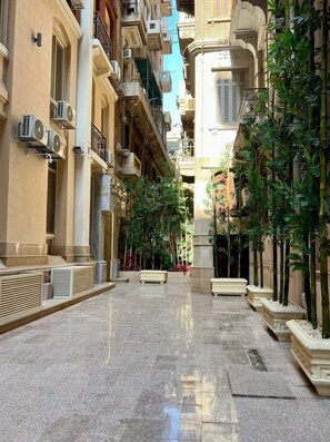Une rue étroite en milieu urbain, bordée de bâtiments des deux côtés, avec des unités de climatisation et des jardinières plantées d’arbres.