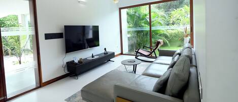 Una sala de estar moderna con un televisor de pantalla plana grande, un sofá gris, una mesita de centro pequeña y un balcón con vistas a zonas verdes.