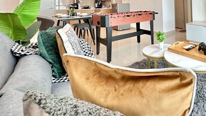 A modern living room with a sofa, a coffee table, and a foosball table. There is a kitchen in the background with cabinets and a window.