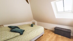 Un dormitorio con cama, almohadas, un banco, una ventana y un techo inclinado.