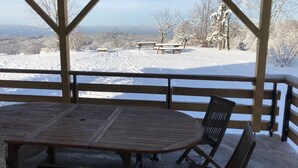 Een overdekt buitengebied met een houten tafel en banken, met uitzicht op een besneeuwd landschap met bomen en verre bergen.