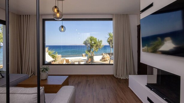 A modern living room with a large window overlooking a beach, a flat-screen TV, and a minimalist design. - Unforgettable Tinos Beach house III (Tinos)