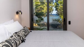 A bedroom with a bed, a nightstand, a wall-mounted lamp, and a sliding glass door leading to a view of a beach and ocean. - Unforgettable Tinos Beach house III (Tinos)