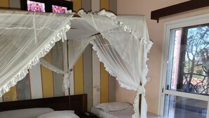 A room with two beds, one with a canopy and one without. There is a window with a view of trees and a balcony. - CENTRAL HOTEL  (Antsiranana)