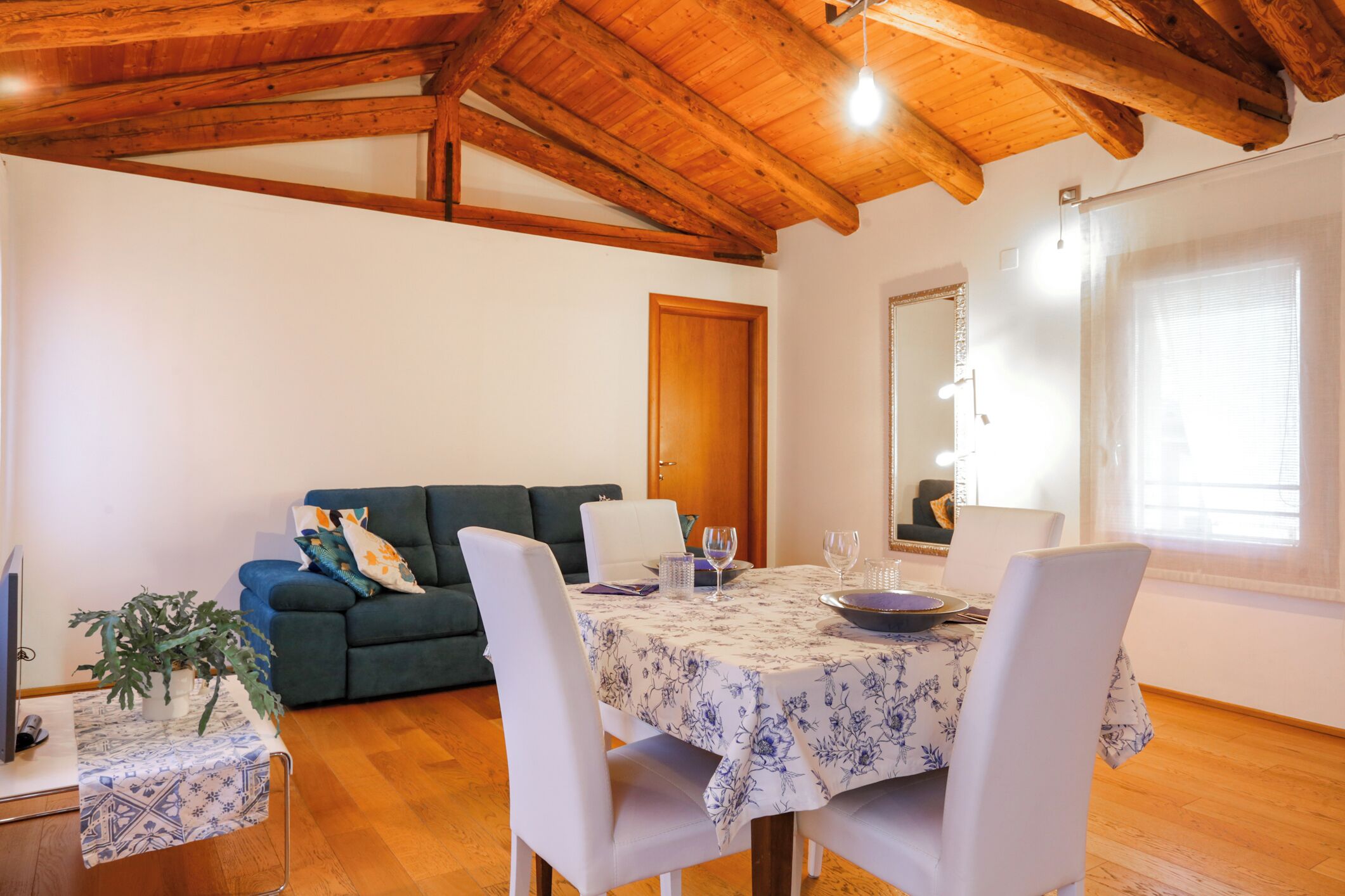 A dining area with a table set for two, a green sofa, wooden ceiling beams, and a large window.
