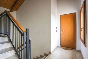 Un escalier avec une rampe en bois et des balustrades noires, une porte en bois avec un paillasson, et un sol carrelé.