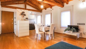 Amplia sala de estar con techo de madera, una mesa de comedor con sillas, una cocina al fondo y un televisor sobre un mueble.