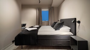 A modern hotel room with a large bed, a desk, and a view of the city through the window.