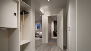 A hallway with a wooden floor, a coat rack, and a door leading to a room with a bed and a window.