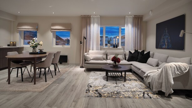 A modern living room with a dining area, beige sofas, a coffee table, and a view of the cityscape through large windows.