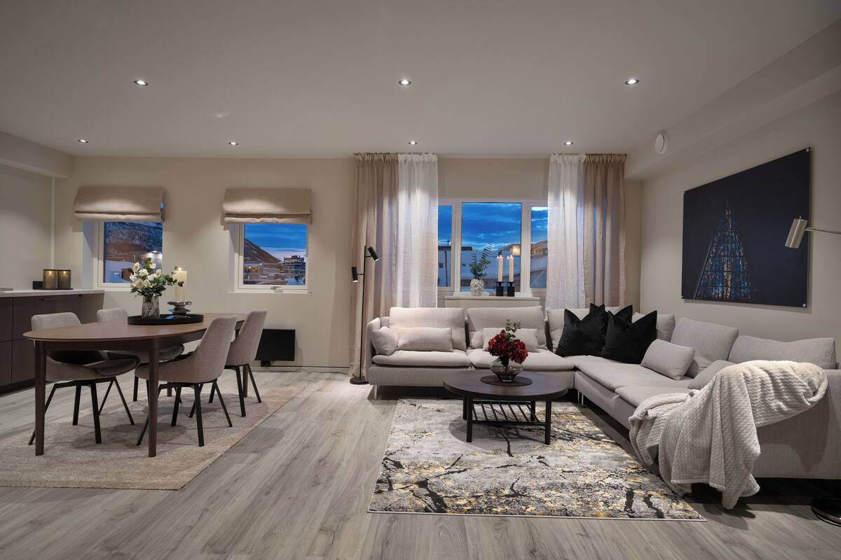 A modern living room with a dining area, beige sofas, a coffee table, and a view of the cityscape through large windows.