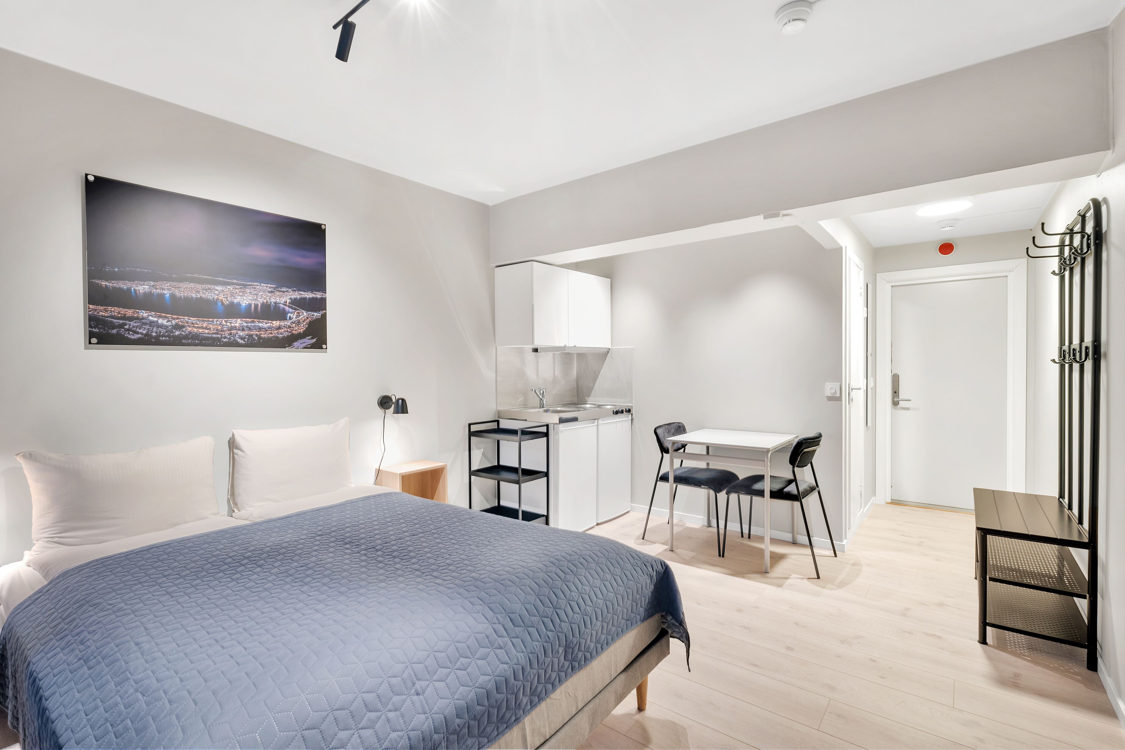 A modern hotel room with a bed, a desk, a chair, and a kitchenette.
