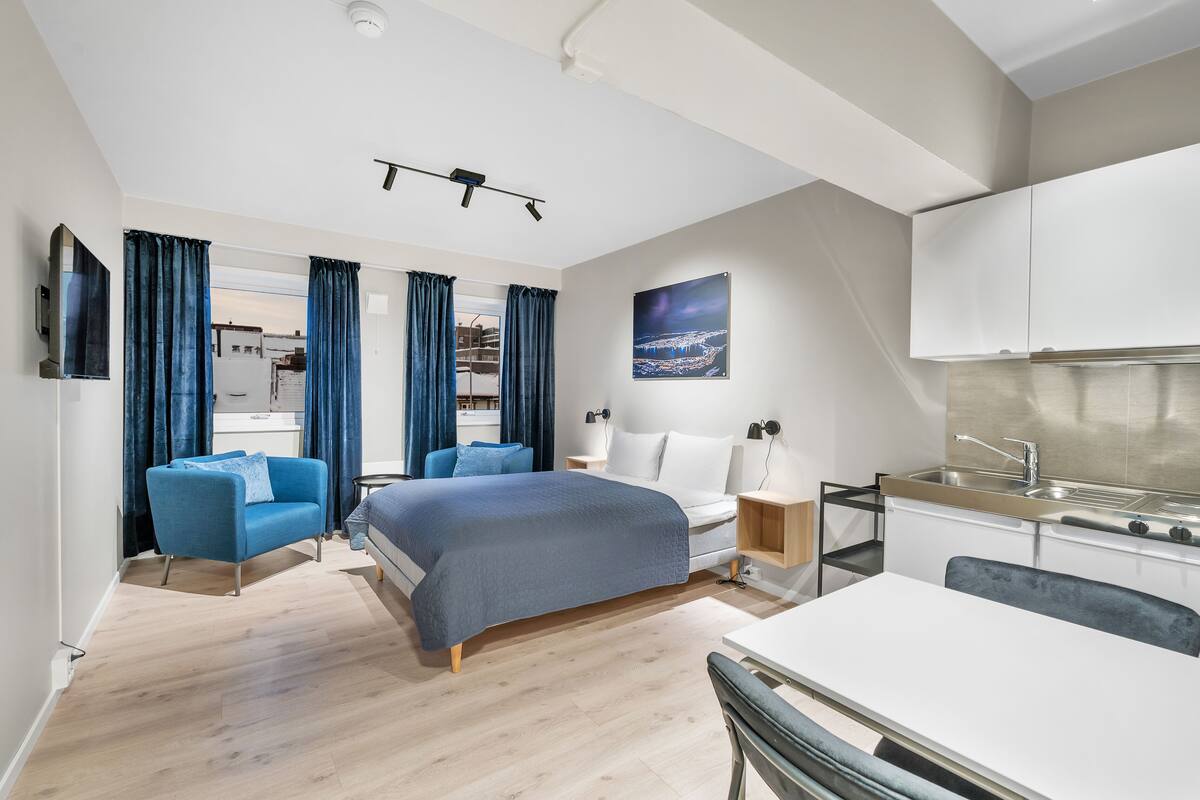 A modern hotel room with a bed, a desk, a kitchenette, and a view of the cityscape.