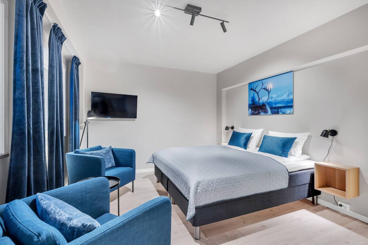 A modern bedroom with a bed, two blue armchairs, a TV, and a painting on the wall.