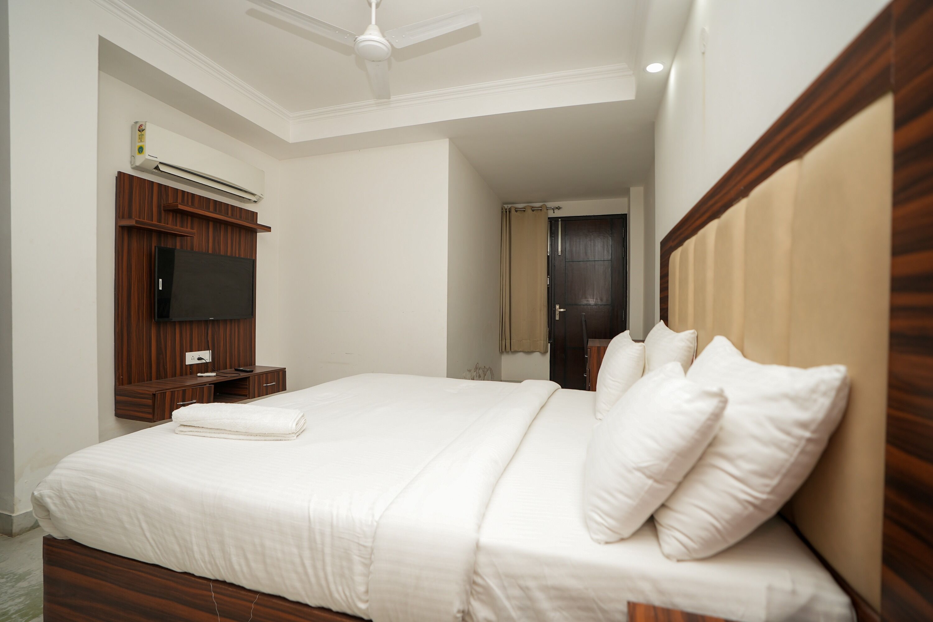 A hotel room with a large bed, wooden headboard, a flat-screen TV, a bedside table, and an air conditioning unit.